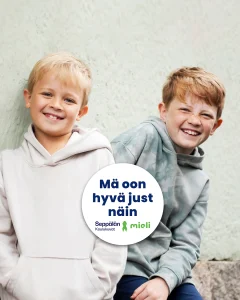 Kaksi hymyilevää lasta istumassa vierekkäin. Kuvassa teksti ”Mä oon hyvä just näin” sekä Seppälän Koulukuvien ja MIELI ry:n logot
